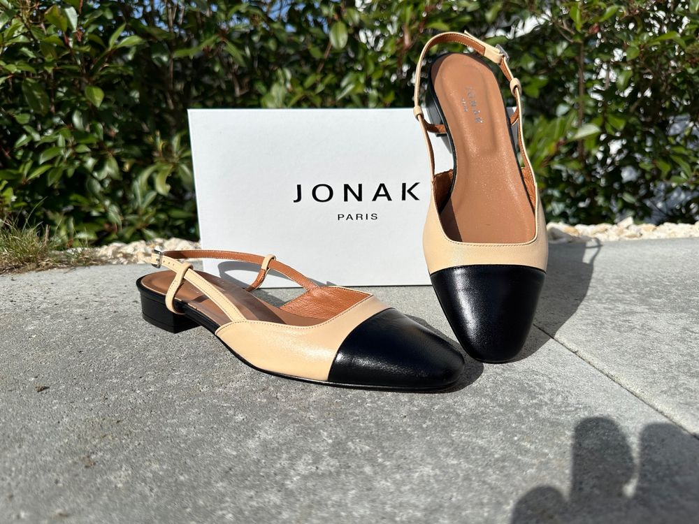 A pair of chic slingback flats in beige leather with contrasting black cap toes is displayed outdoors on a sunlit stone surface. Behind them sits a white box with the “Jonak Paris” logo, emphasizing the shoes’ elegant, minimalist French design.