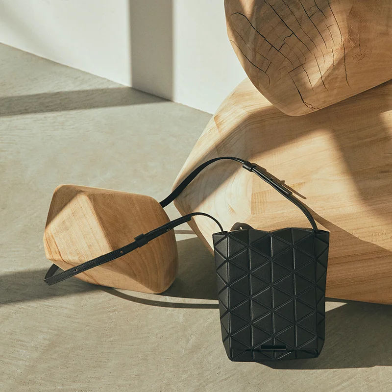 A small black BAO BAO ISSEY MIYAKE crossbody bag with a geometric triangular panel design rests on the floor, leaning against large sculptural wooden blocks in a sunlit minimalist setting.