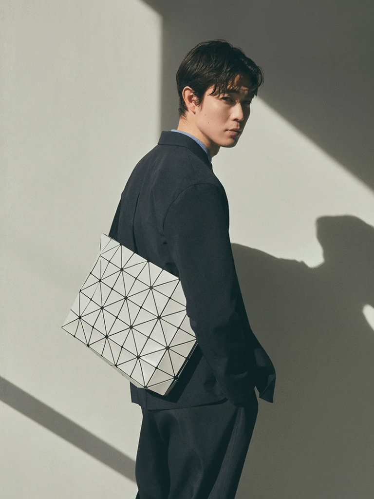 A man dressed in a dark suit stands in partial sunlight against a minimalist wall, carrying a white BAO BAO ISSEY MIYAKE bag with a signature triangular panel design over his shoulder.