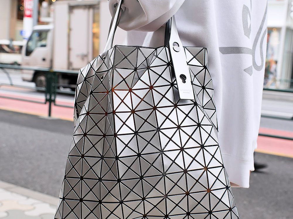 A person wearing a white oversized sweatshirt carries a metallic silver BAO BAO ISSEY MIYAKE tote bag with a geometric triangular panel design, photographed on a city street.