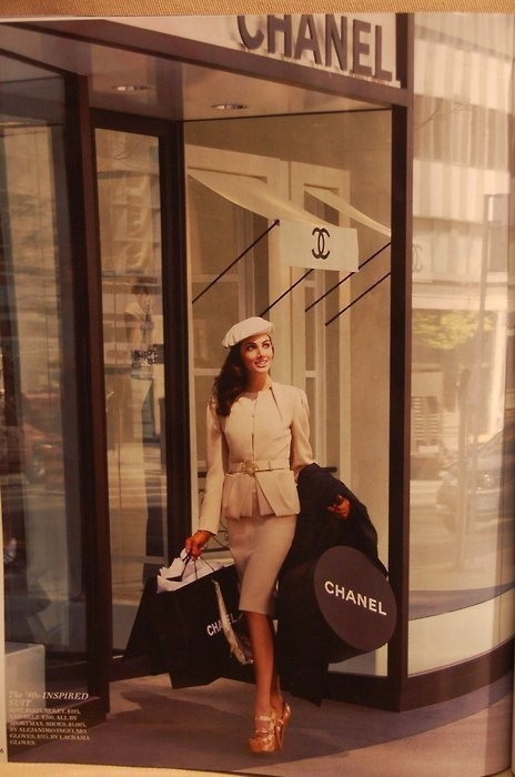 A stylish woman steps out of a Chanel boutique carrying two large round Chanel shopping bags. She wears an elegant beige ensemble with a belted jacket, a fitted pencil skirt, a white beret, and gold platform heels. The glass storefront behind her displays the Chanel logo and a chic awning, evoking a sophisticated Parisian shopping scene.