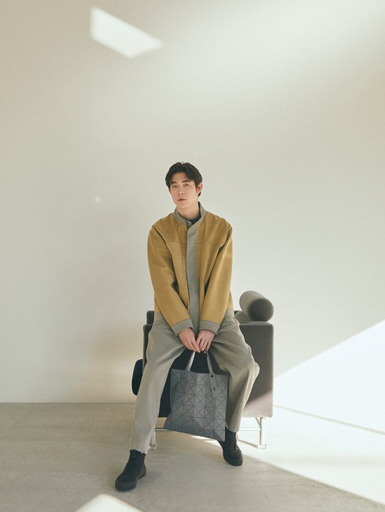 A man sits on a modern grey armchair in a minimal, sunlit room. He wears a mustard-yellow jacket draped over a light grey outfit, paired with dark shoes. He holds a geometric-patterned grey tote bag between his legs, looking directly at the camera with a calm expression.