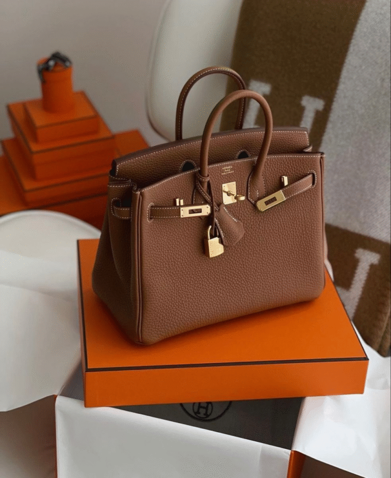 A brown leather Hermès Birkin bag with gold hardware sits atop an orange Hermès box. In the background are neatly stacked orange Hermès packaging and a brown Hermès throw blanket draped over a white chair.