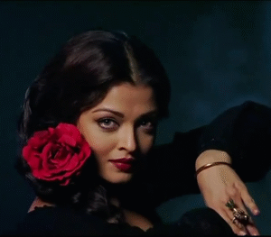 From the film Guzaarish, this visual captures a character styled in deep black with classic red lips, delicate jewelry, and a dramatic rose accessory, embodying the romance and intensity of the scene’s aesthetic.