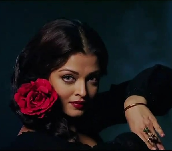 From the film Guzaarish, this visual captures a character styled in deep black with classic red lips, delicate jewelry, and a dramatic rose accessory, embodying the romance and intensity of the scene’s aesthetic.