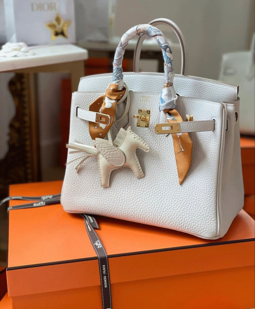 A white Hermès Birkin bag with gold hardware, styled with a horse-shaped bag charm and silk Twilly scarves wrapped around the handles, rests on an orange Hermès gift box. Dior packaging and additional Hermès boxes are visible in the background.