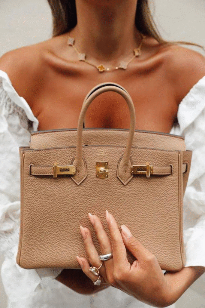 A woman in an off-shoulder white blouse holds a tan Hermès Birkin bag with gold hardware close to her chest. She wears a delicate clover necklace and rings, with manicured nails accentuating the luxury accessory.