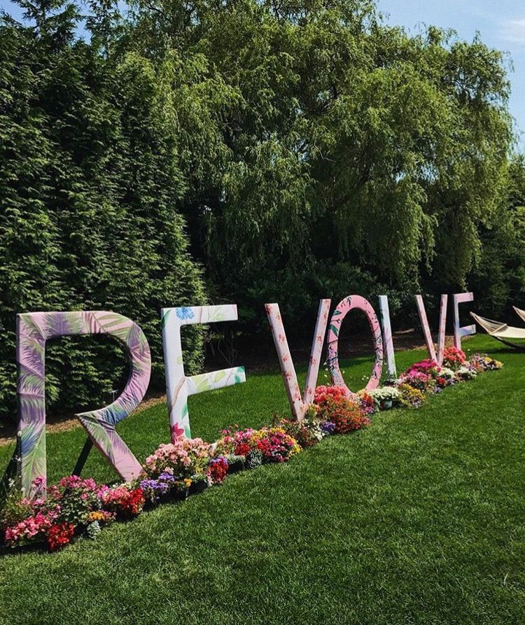 Large freestanding letters spelling “REVOLVE” decorated with colorful floral and tropical patterns, displayed outdoors on a lush green lawn. Bright flowers are arranged along the base of the letters, and tall trees in the background create a vibrant, summery setting.
